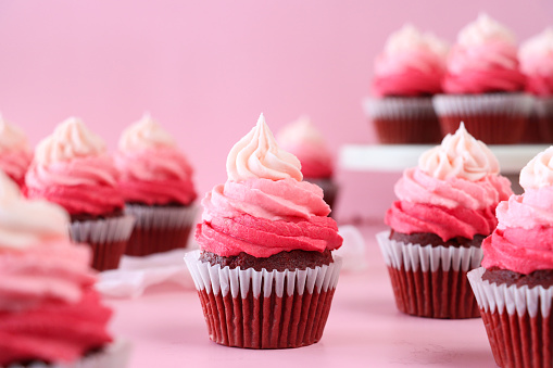 Stock photo showing close-up view of a batch of freshly baked, homemade, red velvet cupcakes in paper cake cases on and surrounding a cake stand. The cup cakes have been decorated with swirls of ombre effect pink piped icing.  Valentine's Day and romance concept.