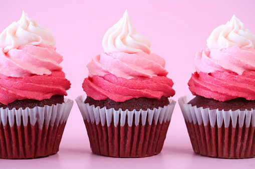 Stock photo showing close-up view of a row of three, freshly baked, homemade, red velvet cupcakes in paper cake cases. The cup cakes have been decorated with swirls of ombre effect pink piped icing.  Valentine's Day and romance concept.