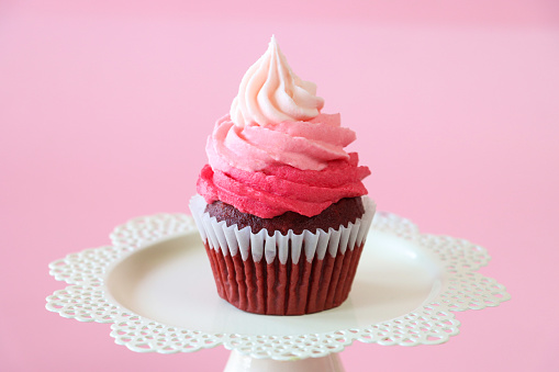 Stock photo showing close-up view of a single freshly baked, homemade, red velvet cupcake in paper cake case on a white, cake stand. The cup cake has been decorated with a swirl of ombre effect pink piped icing.  Valentine's Day and romance concept.