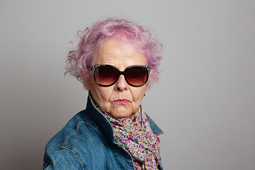 Senior woman with dyed pink hair, wearing sunglasses,  jeans jacket and scarf with floral pattern, cool attitude