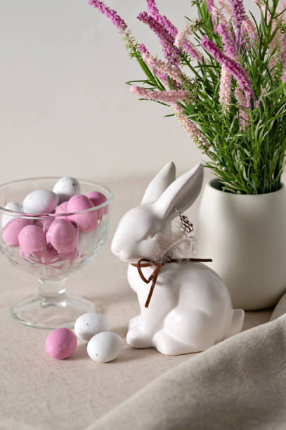 istock Lifestyle Easter holiday still life, home interior decor. Rabbit figurine, pink and white egg candies, flowers in vase on neutral beige linen tablecloth background, soft natural light, selective focus 1988138035