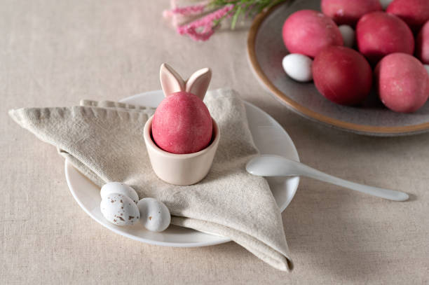 istock Easter holiday table setting, pink colored egg in cup with bunny ears, on plate with napkin, spoon and candies, on neutral beige linen tablecloth background, soft natural sunlight, selective focus 1988138611