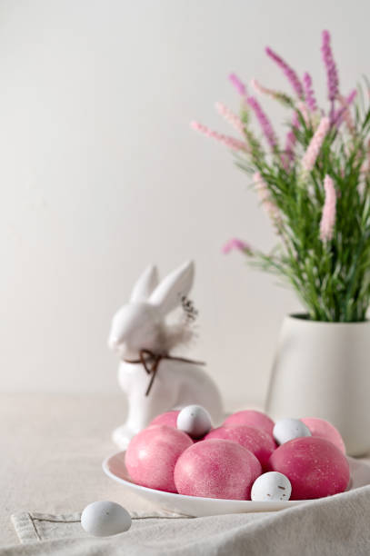 istock Traditional Easter food and table setting decor, pink dyed eggs on plate, rabbit figurine, spring flowers in vase on beige linen tablecloth, blurred background, soft natural light, selective focus 1988140253