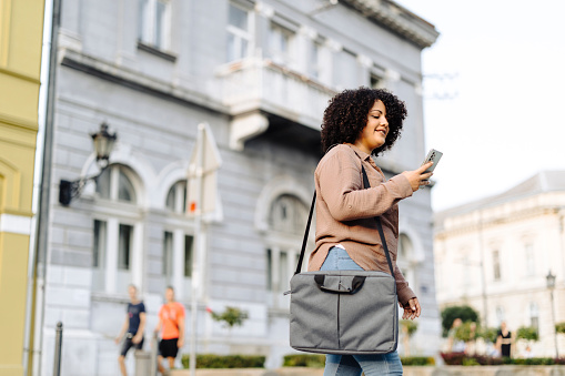 Shot of young African-American businesswoman walking through the city holding her laptop bag, using phone and looking away.