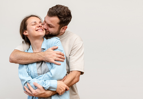 Happy young woman in the arms of her husband in front of light grey background win studio. Man hugs woman.