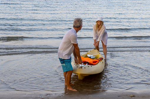 Mature couple pull sea kayak onto beach at golden hour