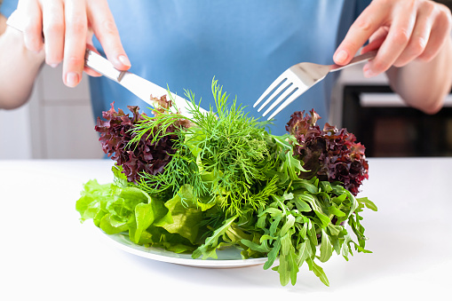 The concept of a healthy, balanced, dietary food. Girl with knife and fork eating fresh vitamin green salad with arugula, dill and lettuce. Close-up.