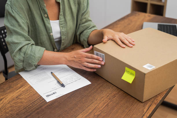 istock online shopping refund, a woman places a barcode sticker on a cardboard parcel for return. 1988306277