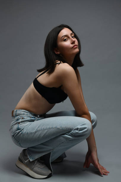 istock Female Squatting Down While Wearing Jeans And Black Bra On Grey Background 1988332229