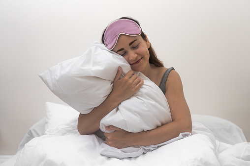 A captivating young woman, sporting a sleep mask, hugs a soft white pillow, exuding morning contentment in her bedroom.
