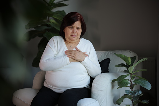Heart attack, women with chest pain suffering at home, health problems concept. Young women with eyes closed holding his chest in discomfort, suffering from chest pain while sitting on sofa at home. Elderly and health issues concept