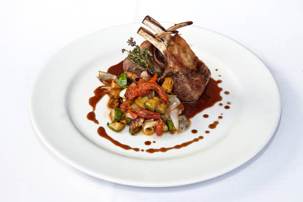 Roasted rack of lamb or rack of veal accompanied with vegetables and sauce, served on white plate over white background