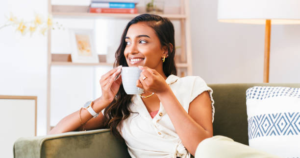 istock Smile, thinking and Indian woman on sofa for coffee break, daydreaming or quiet reflection in her home. Idea, inspiration and calm female person with insight, memory or peaceful moment in living room 1988358846