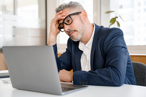 Fatigued mature businessman using laptop for working, man in formal wear feeling tired and burnout, sitting at the desk in modern office, doubting and thinking