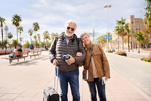 Smiling Caucasian mature tourist couple standing posing looking at the camera in the street with their luggage. Husband and wife enjoying their retirees holidays wearing winter clothes on sunny day