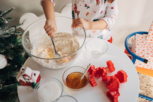 The child is using a spoon to mix the ingredients for cookies. The toddler is adding , and mixing the flour, eggs, honey and sugar in the glass bowl, and making gingerbread cookies.