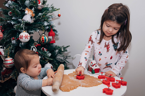The three-year old girl and her baby sister are making Christmas cookies. The children are using plastic module to cut the dough and making gingerbread cookies.