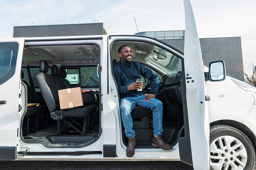 A happy delivery driver is sitting in the open door of his white van, holding a cup and enjoying a break in Ljubljana, Slovenia