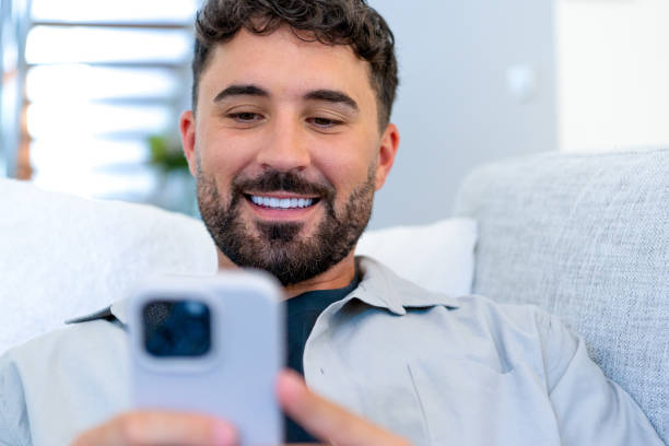 istock Happy smiling young man relaxing sitting and reclining on the sofa at home. He is using a mobile phone 1988490679