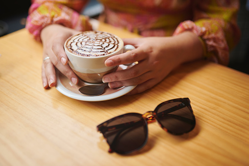 Hands, coffee cup and woman at cafe closeup with sunglasses for break, relax or chilling. Latte art, restaurant or fingers with beverage, liquid or drink for caffeine satisfaction on travel trip