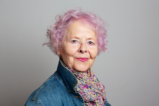 Senior woman with dyed pink hair, wearing jeans jacket and scarf with floral pattern, smiling at camera