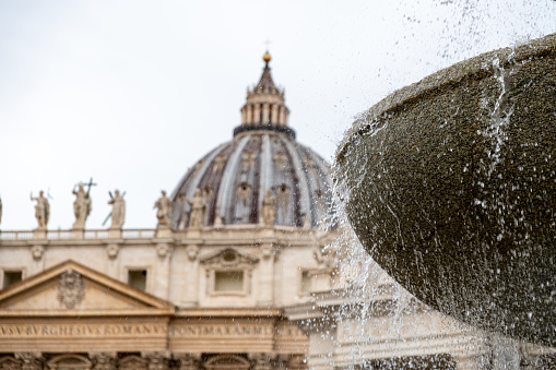 Roma, Latium - Italy - 11-26-2022: Water cascades down a fountain with St. Peter's Basilica's dome majestically poised in the soft-focus background