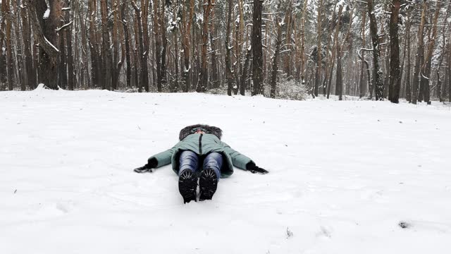 Happy woman lying on snowy lawn at forest and making angel. Young girl playing in snow and enjoying wintertime. Cheerful lady having fun outdoor. Concept of a winter vacation. Close up Slow mo