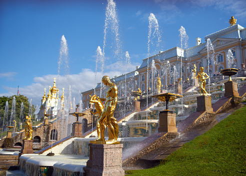 Saint Petersburg, Russia - June 09, 2023: Golden monuments with fountains and Grand Palace in Peterhof park near St. Petersburg with blue sky. Built in 18th century laid out on the orders of Peter the Great. The palace-ensemble with fountains, gardens and monuments is recognized as a UNESCO World Heritage Site.