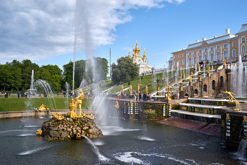 Saint Petersburg, Russia - June 09, 2023: Golden monuments with fountains and Grand Palace in Peterhof park near St. Petersburg with blue cloudy sky. Built in 18th century laid out on the orders of Peter the Great. The palace-ensemble with fountains, gardens and monuments is recognized as a UNESCO World Heritage Site.