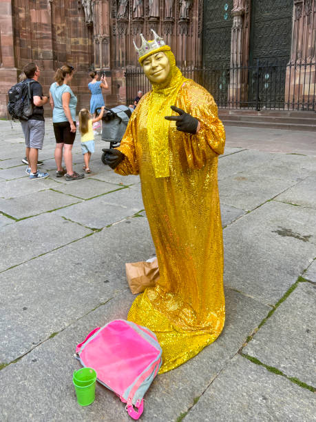 Strasbourg, France, Europe, July 20, 2023, Human Mime Give the Peace Sign in the town square. She is standing in front of the Strasbourg cathedral and is dressed in all gold with gold face paint. She is wearing a crown.