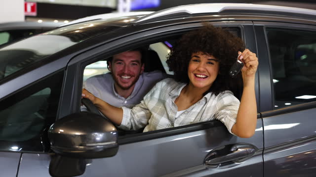 Excited young couple sitting in their new car and then facing the camera smiling very cheerfully while woman holds the key