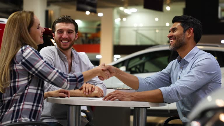 Happy Latin American couple closing a deal at the car dealership handshaking a friendly salesman