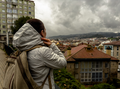 A solo traveler stands with her back to the camera, gazing over a sprawling cityscape from a vantage point. Her hooded jacket and backpack suggest a readiness for exploration, despite the overcast sky above. There's a quiet contemplation in her posture, a story of wanderlust and the reflective journey of traveling alone.