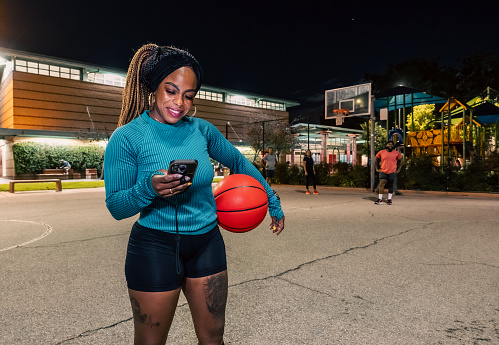 A medium wide portrait of a Black woman holding a basketball on her side and looking at her smartphone while her friends watch on from the background.