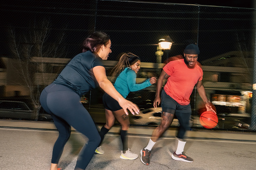 A blurred action shot of a Black man dribbling the basketball away from his friends, a Hispanic woman and Black woman, while playing on a concrete court in Los Angeles at night.