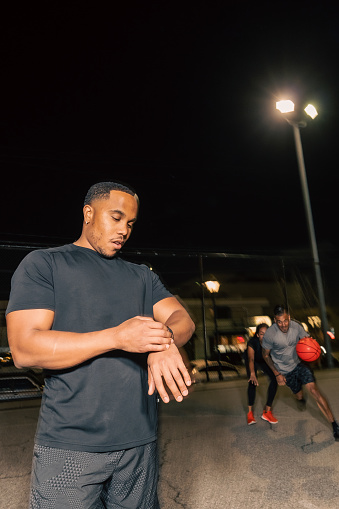 A portrait shot of a multiracial man looking at his watch while his friends play basketball behind him on a concrete court in Los Angeles at night.