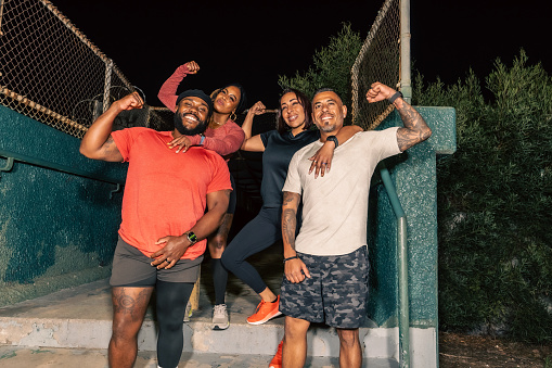 Four friends, two Black people and two Hispanic people, posing and flexing their arms for the camera after working out at night.