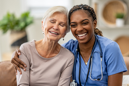 A Home Care nurse sits with a senior resident as they pose for a portrait together.  The nurse is wearing blue scrubs and the senior is dressed comfortably as they both smile.