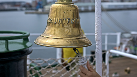 a hand reaching for the rope of a ship's bell, poised to ring out in clear, resonant tones. The bell, inscribed with the word CUAUHTEMOC, shines with the patina of age and use, reflecting its storied past aboard the vessel. A glimpse of the deck's white railing and the blurred harbor in the distance create a backdrop that evokes the timeless ritual of signaling the watch change or marking significant moments at sea.