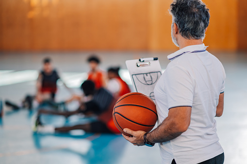 Rear view of a senior basketball trainer standing on court with clipboard and basketball in his hands and looking at his team in blurry background. An old coach is training his basketball team.