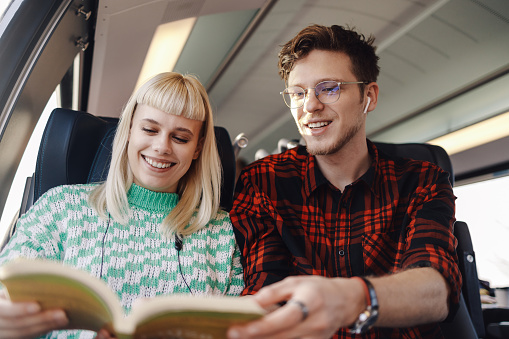 Portrait of a young passengers sitting in a train and reading a book together while traveling. A hipster couple is commuting by public transportation, reading a book and smiling at it.Couple traveling