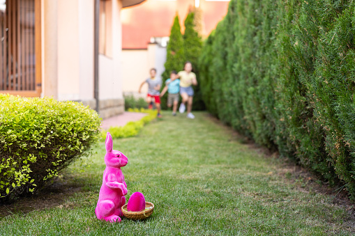 Excited children are running towards a pink Easter rabbit sitting on the ground in the foreground outside on a sunny day in a garden