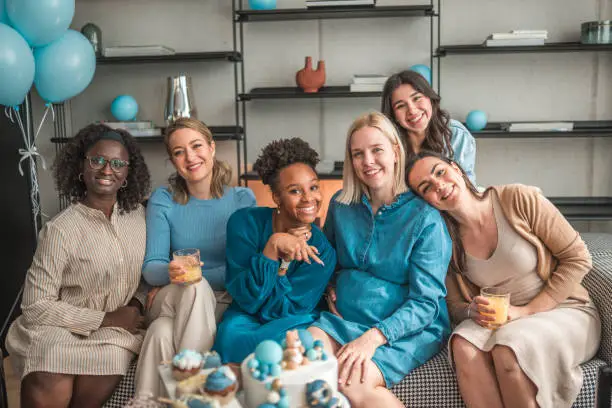 Cheerful girlfriends come together in a beautifully decorated living room for a baby shower, gifting their blonde pregnant friend with love and presents.