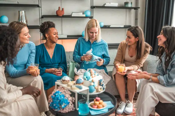A group of happy girlfriends sitting on a sofa, surrounded by balloons and sweets, while gifting their Caucasian pregnant blonde friend in a baby shower celebration.
