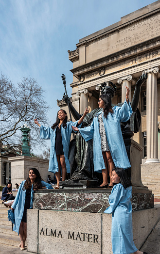 South Asian and East Asian young women Columbia graduates mark their graduation by standing at the “Alma Mater” statue on the steps of Low Library at Columbia University.

The statue’s sculptor was Daniel Chester French (1850-1931,) who, among other famous works, designed the Lincoln statue at the Lincoln Memorial in Washington DC.