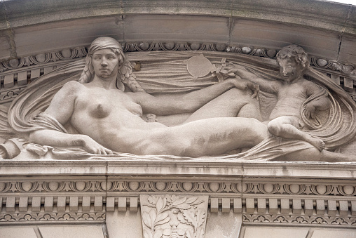 A classical reclining female nude figure graces the pediment over the entrance to the Frick Mason on E. 70th Street. On the right side, a cupid holds a mirror for the beauty. \n\nSherry Edmundson Fry (1879-1966) designed the sculpture, which was carved into the marble stone by Attilio Piccirilli ((1866-1945.) \n\nFry’s model was Audrey Munson. Munson posed for more than a dozen important statues, sculptures, and friezes, most of which still stand to this day across Manhattan and Brooklyn. She is now often called “America’s First Supermodel.”