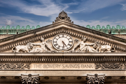 Completed in 1895, the Bowery Savings Bank Building at 130 Bowery was designed by Stanford White (1853-1906.) The building was designed in a Greco-Roman classical style, with Corinthian columns and a triangular pediment on the front facade. The pediment was sculpted by Frederick William MacMonnies (1863-1937.) and features two classically draped female figures taming a lion each, situated on either side of a large clock. Assorted fruits are also carved into the pediment as an allegory for bountiful wealth. 

Bowery Savings Bank at one time had 35 branches across New York City, but by 1985, with dwindling assets, it was sold to another financial institution. The bank went through several more sales until the assets were sold to Capital One Bank in 2007. 

The building is a New York City designated landmark, and is currently used as a high-end venue.