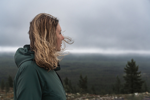 Side face view of Caucasian woman with blond hair blowing in wind in nordic nature, female hiker standing on mountain, in mind looking away over the forest and stormy clouds in the background