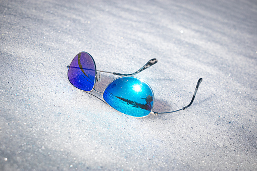 Photo of blue-lense sunglasses with reflection of the sun and landscape on an angle on a patch of snow in the Sierra Nevada mountains.