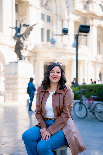 Happy young hispanic woman while sitting in front of blurred Bellas Fine Arts Palace in Mexico City in daylight enjoying location and wearing fancy clothes